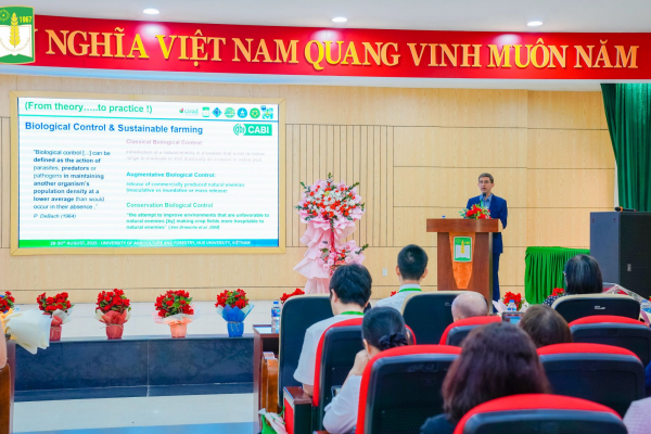 Arnaud Costa of CABI speaks at a podium during ICBPSA25 in Huế, with a large screen behind him showing slides on biological control and sustainable farming, while the audience listens from red-seated rows.