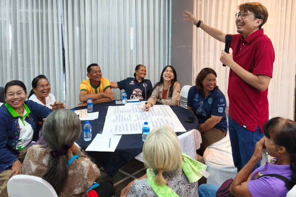 Discussion de groupe avec des agriculteurs impliqués dans le projet « Khok Nong Na » assis autour d'une table en train de rire avec le coordinateur qui tient le micro.