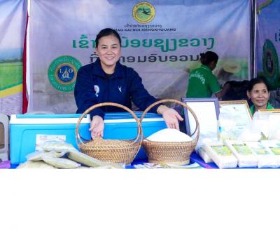 Une femme se tient devant un &eacute;tal de march&eacute; au Laos, pr&eacute;sentant des paniers de riz Khao Kai Noi ainsi que des produits &agrave; base de riz emball&eacute;s et des documents de certification, avec une banderole identifiant le produit derri&egrave;re elle.