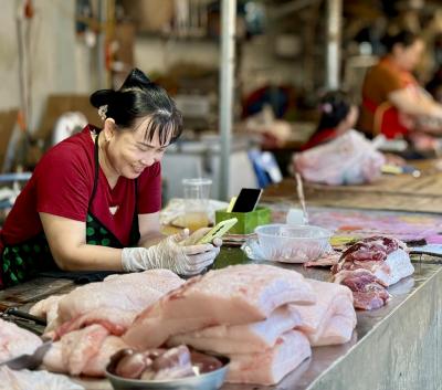 A pork vendor wearing gloves smiles while using her phone at a traditional market stall in Dien Bien province, Vietnam.